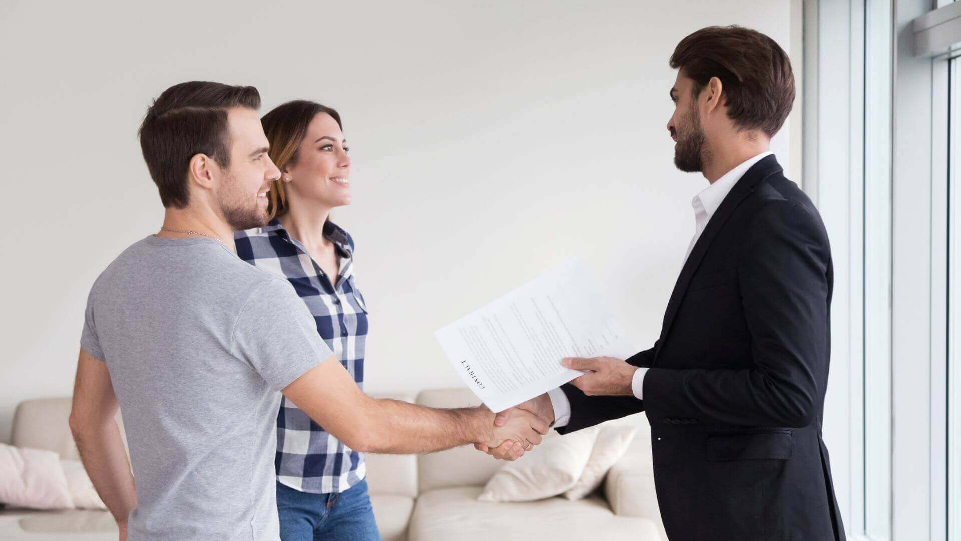A landlord in a black suit with a beard holds a lease agreement while shaking hands with two new, smiling tenants.