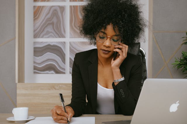 woman in a black blazer on a phone call at a desk with a notepad and an open laptop